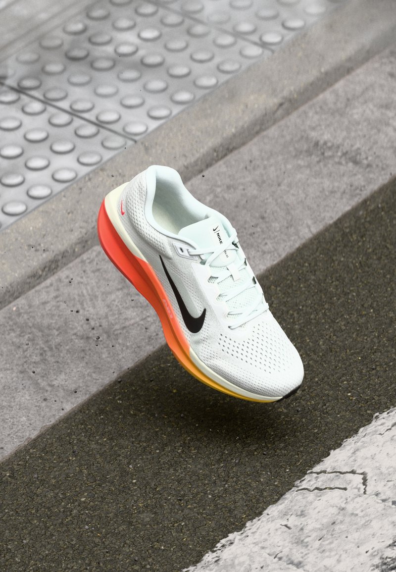 Chaussure de course Nike blanche avec swoosh noir et semelle dégradée orange sur l’asphalte près d’un pavé tactile et d’un passage piéton.
