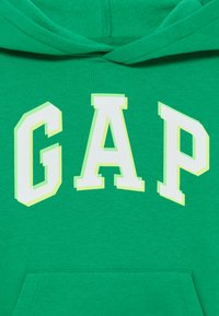 Grønn hettegenser med en stor, hvit "GAP"-logo med lys grønne konturer. Myk tekstur, pullover-design med en framlomme.
