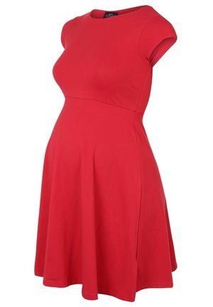 Kvinde i knælængde rød kjole og sorte snørelåste ankelstøvler med hæl, stående mod en ensartet lys baggrund.