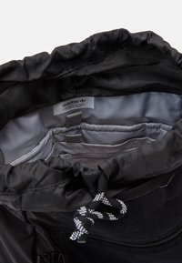 Černý sáček na stažení s hladkým vnitřkem. Obsahuje značkovou etiketu, dvě přihrádky a kontrastní bílé detaily na šňůrkách.