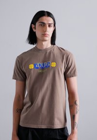 Pruun puuvillane T-särk ümmarguse kaelusega. Ees on sinine ja kollane "KIDSUPER" logo koos lillemustritega. Lühikesed varrukad.