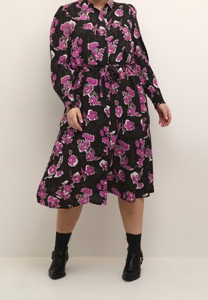 Zwarte midi-jurk met roze en witte bloemenprint, lange mouwen, knopen aan de voorkant, tailleband, gedragen met zwarte enkellaarsjes.
