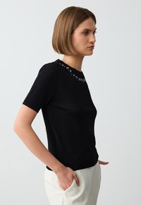 Schwarzes kurzärmliges Strickoberteil mit Rundhalsausschnitt, der mit dekorativen Steinen verziert ist. Es zeigt eine figurnahe Silhouette und eine glatte Textur.