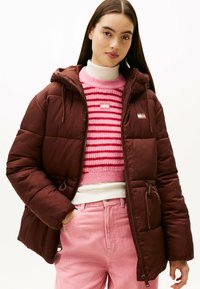 Bruine gewatteerde jas met een capuchon, voorzien van een rits en zijzakken. Gedorven over een roze en witte gestreepte trui en roze corduroy broek.