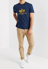 Námořnická modrá tričko s žlutým logem "Alpha Industries", kombinované s béžovými chinos kalhotami a bílými teniskami. Ležérní střih, krátké rukávy, kulatý výstřih.