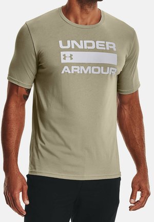 Mand iført en lyse olivengrøn Under Armour T-shirt med hvidt logo og tekst på brystet, kombineret med sorte bukser.