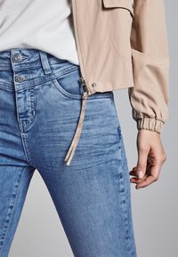 Chaqueta bomber beige claro sobre una camiseta blanca con jeans azules. Los jeans tienen un brillo sutil y un cierre con cremallera con un tirador beige.