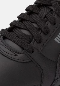 Mustat urheilujalkineet, joissa on teksturoidusta synteettisestä nahasta valmistettu päällinen, varustettuna litteillä mustilla kengännauhoilla ja logoyksityiskohdalla. Lähikuva kengännauha-alueesta ja materiaalista.