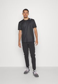 Melnas acu auduma sporta t-krekls ar īsām piedurknēm un trim svītrām augšējā kreisajā stūrī. Apvienots ar iegurņa piegriezuma melnajiem biksēm un pelēkiem sporta apaviem.