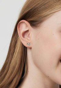 Srebrne kolczyki w kształcie kokardy, małe, o gładkiej fakturze i minimalistycznym wzornictwie, noszone na uchu.