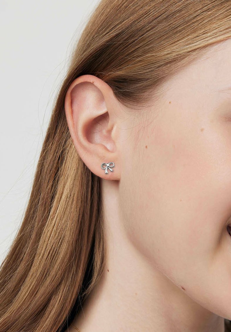 Srebrne kolczyki w kształcie kokardy, małe, o gładkiej fakturze i minimalistycznym wzornictwie, noszone na uchu.