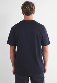 Homme avec des cheveux ondulés châtain clair portant un t-shirt uni bleu marine et un pantalon sombre, se tenant contre un fond blanc, tourné le dos à l'appareil photo.