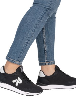 Personne portant un jean bleu et des baskets noires avec des semelles blanches et un logo, debout avec un pied légèrement levé.