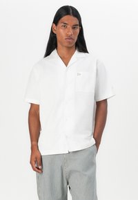 Camicia bianca a maniche corte con bottoni e taschino, con un logo sottile, abbinata a pantaloni corti grigi a righe. Tessuto dalla superficie liscia.