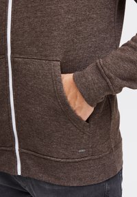 Brun zip-up hoodie lavet af tekstureret stof, med en hvid lynlås, frontlomme og et lille mærke på den nederste side.