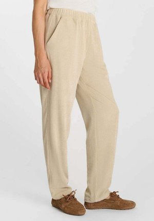 Persona che indossa pantaloni beige texturizzati con vita elastica e scarpe in mocassino di camoscio marrone, in piedi su sfondo bianco.
