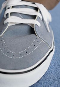 Grijze suède sneaker met witte veters, voorzien van geperforeerde details, witte tussenzool en een zwarte accent langs de rand van de zool.