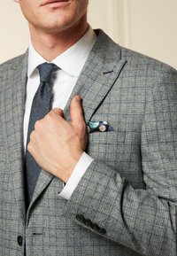 Szary garnitur w wzór z teksturowanym wykończeniem, w zestawieniu z białą koszulą elegancką i ciemnym krawatem. Poszetka ma okrągłe niebieskie i różowe wzory.