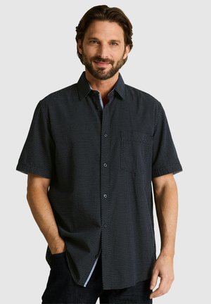 Man with beard wearing a short-sleeve, dark button-up shirt with small white dots, standing with one hand in pocket against plain background.
