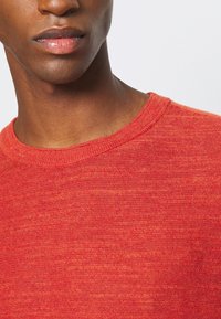 Pull en tricot rouge avec un col rond et un tissu texturé, mettant en valeur un mélange de nuances tonales.