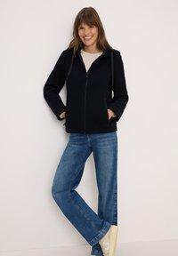 Veste en polaire marine à zip avec capuche, deux poches latérales, associée à un jean en denim bleu clair et des baskets beiges. Design simple et décontracté.