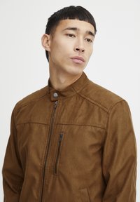 Veste en daim marron avec col montant, fermeture éclair sur le devant et deux poches avant, le tout avec une texture lisse et mate. Détails de couture simples.