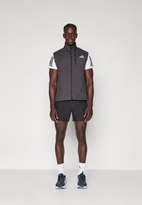 Schwarze gepolsterte Weste mit hohem Kragen, Frontreißverschluss und kleinem Logo. Getragen über einem weißen T-Shirt, kombiniert mit schwarzen Shorts und Laufschuhen.