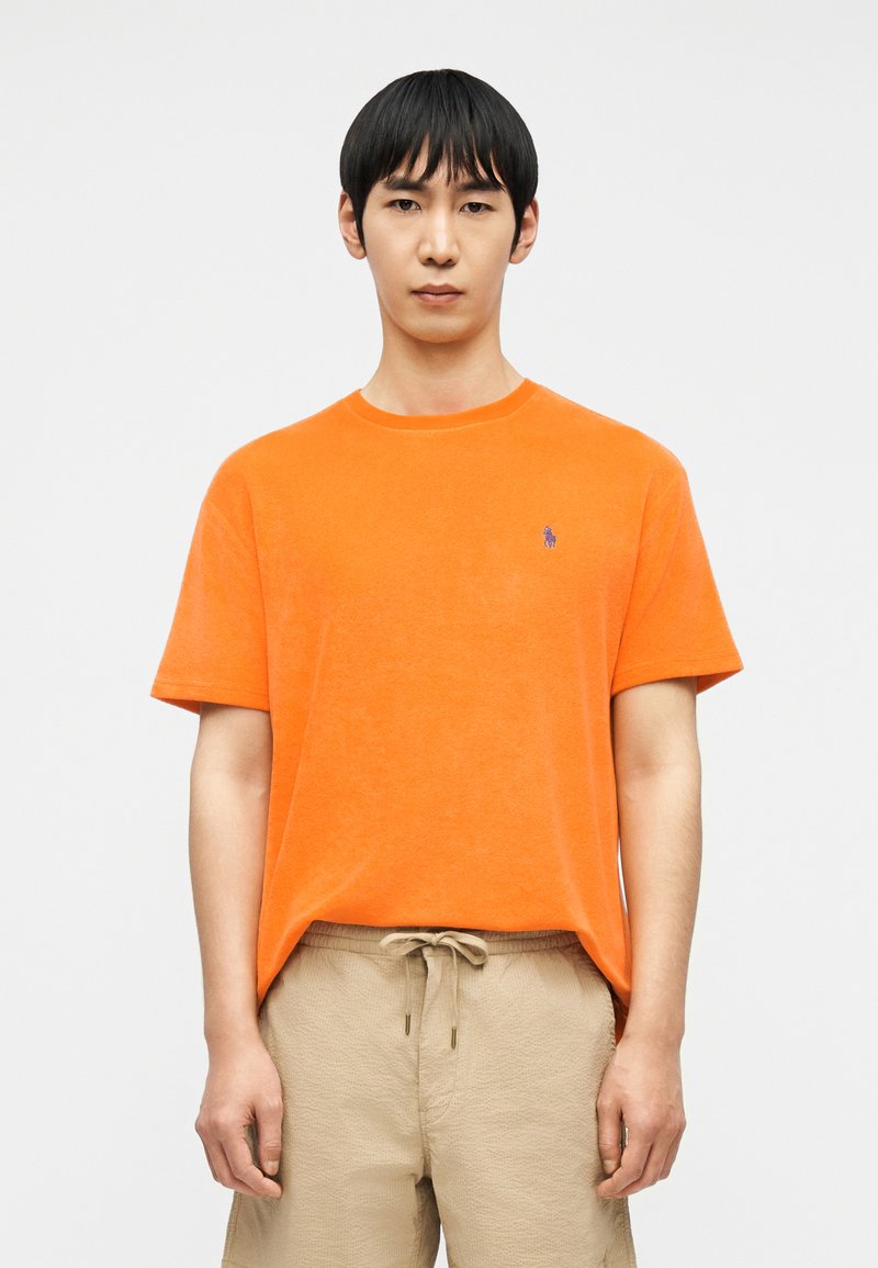 Άνδρας που φοράει φωτεινό πορτοκαλί κοντομάνικο πουκάμισο με μικρό λογότυπο στο στήθος και μπεζ σορτς με κορδόνι, στέκεται μπροστά σε απλό λευκό φόντο.