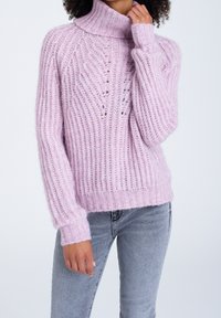 Ljusrosa polotröja i stickad struktur med ribb och utskärningar, visad med grå jeans. Mjuk och mysig look.