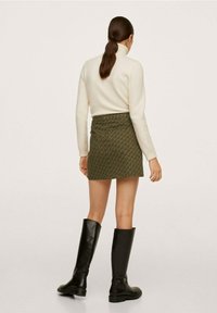 Pull à col roulé crème clair, mini-jupe verte à motifs, et bottes en cuir noires montant jusqu'aux genoux. Mise en avant sur la texture et le contraste des couleurs.