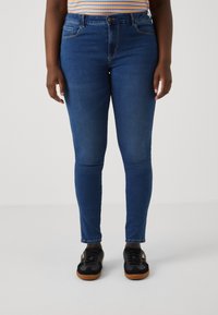 Skinny jeans in dark blue denim, featuring a mid-rise waist, five pockets, and subtle stitching accents. Paired with black sneakers.