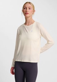 Long-sleeve cream top made of lightweight fabric with a round neckline and small blue logo on the chest, paired with dark leggings.