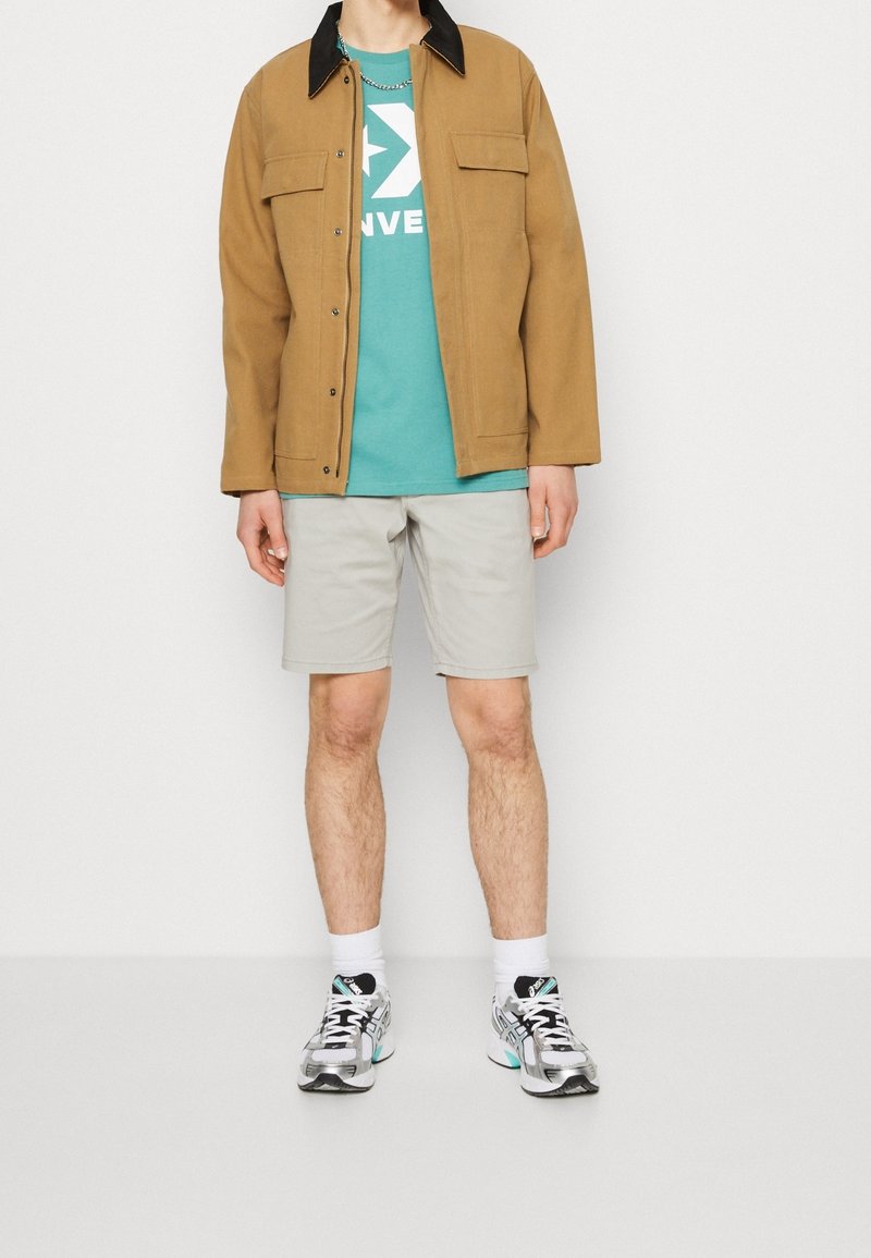 Hombre con chaqueta marrón, camiseta gráfica azul verdoso, pantalones cortos gris claro, calcetines blancos y zapatillas deportivas plateadas, de pie contra un fondo blanco.