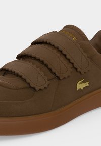 Baskets marron avec trois bandes velcro festonnées, un logo crocodile doré sur le côté, et une semelle en caoutchouc marron assortie.