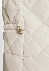 Close-up van beige doorstikte stof met een diamanten patroon en een zilveren cordlock op een gerimpeld gedeelte.