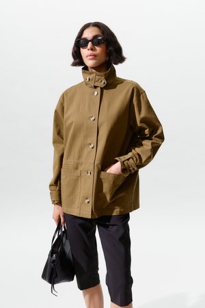 Naine, kes kannab üle suurenenud oliivirohelist jopet, musti põlveni ulatuvaid pükse, musti päikeseprille, hoides käes musta käekotti, seisab valge tausta ees.