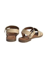Beige leren sandalen met een platte zool, voorzien van kruisbanden en een verstelbare enkelband met een metalen gesp. Bruine inlegzool textuur.