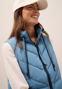 Gilet matelassé bleu clair avec col montant, fermeture éclair sur le devant et cordons de serrage. La partie supérieure présente un tissu lisse, contrastant avec les manches texturées.