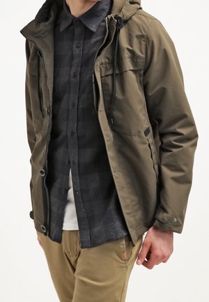 Homme portant une veste vert olive par-dessus une chemise noire à carreaux boutonnée et un pantalon beige, avec une main visible à son côté.