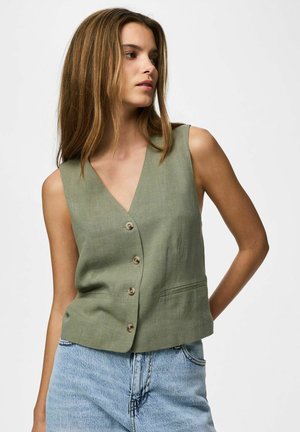 Donna con capelli lisci castano chiaro che indossa un gilet smanicato verde oliva con bottoni e jeans azzurri a vita alta.