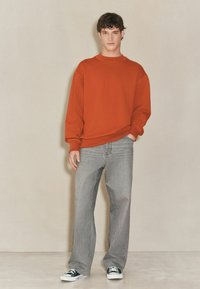 Czerwona bluza z okrągłym dekoltem z opuszczonymi ramionami, zestawiona z szerokimi, jasnoszarymi dżinsami i czarnymi sneakersami. Gładki materiał, casualowy krój.