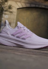 Светлолилав маратонка Adidas Ultraboost с текстурирана подметка и характерен дизайн с три ивици, показана навън върху каменна повърхност.