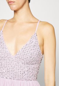 Mujer que lleva un vestido sin mangas de color rosa claro con tirantes finos y un corpiño adornado con lentejuelas plateadas en un patrón de diamantes.
