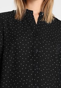 Schwarze button-up Hemd mit einem Kragen, mit weißen Punkten, glatter Textur und einer vorderen Leiste mit gleichmäßig verteilten Knöpfen.