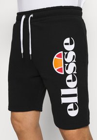 Shorts noirs avec cordons blancs et logo "ellesse" blanc en majuscules disposé verticalement à côté d'un graphique en demi-cercle orange et rouge.