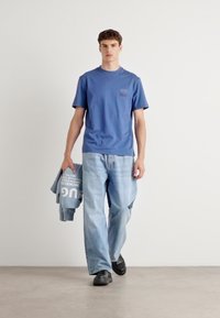 Zila kokvilnas t-kreklu ar īsām piedurknēm un kabatas detaļu, saskaņota ar gaiši ziliem platām biksēm un nesot pelēku džemperi.