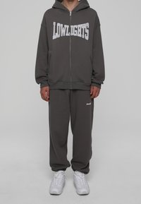 Sudadera con cremallera en gris oscuro con la palabra "LOWLIGHTS" en letras blancas, combinada con pantalones de chándal grises a juego. Ambos artículos tienen un ajuste relajado.