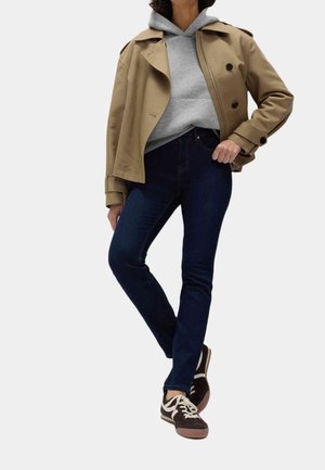Person trägt einen beigefarbenen Trenchcoat über einem grauen Hoodie, dunkelblaue Jeans und braune Sneaker und steht mit einer Hand in der Tasche.