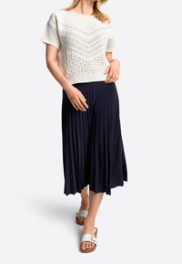 Strikket hvid cropped sweater med et tekstureret mønster, kombineret med et flydende navy plisseret nederdel og hvide slid-sandaler med en guldbeklædning.