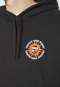 Personne portant un sweat à capuche noir avec un graphisme de soleil orange et un texte blanc « Protégez votre planète » entourant le logo Puma sur la poitrine.